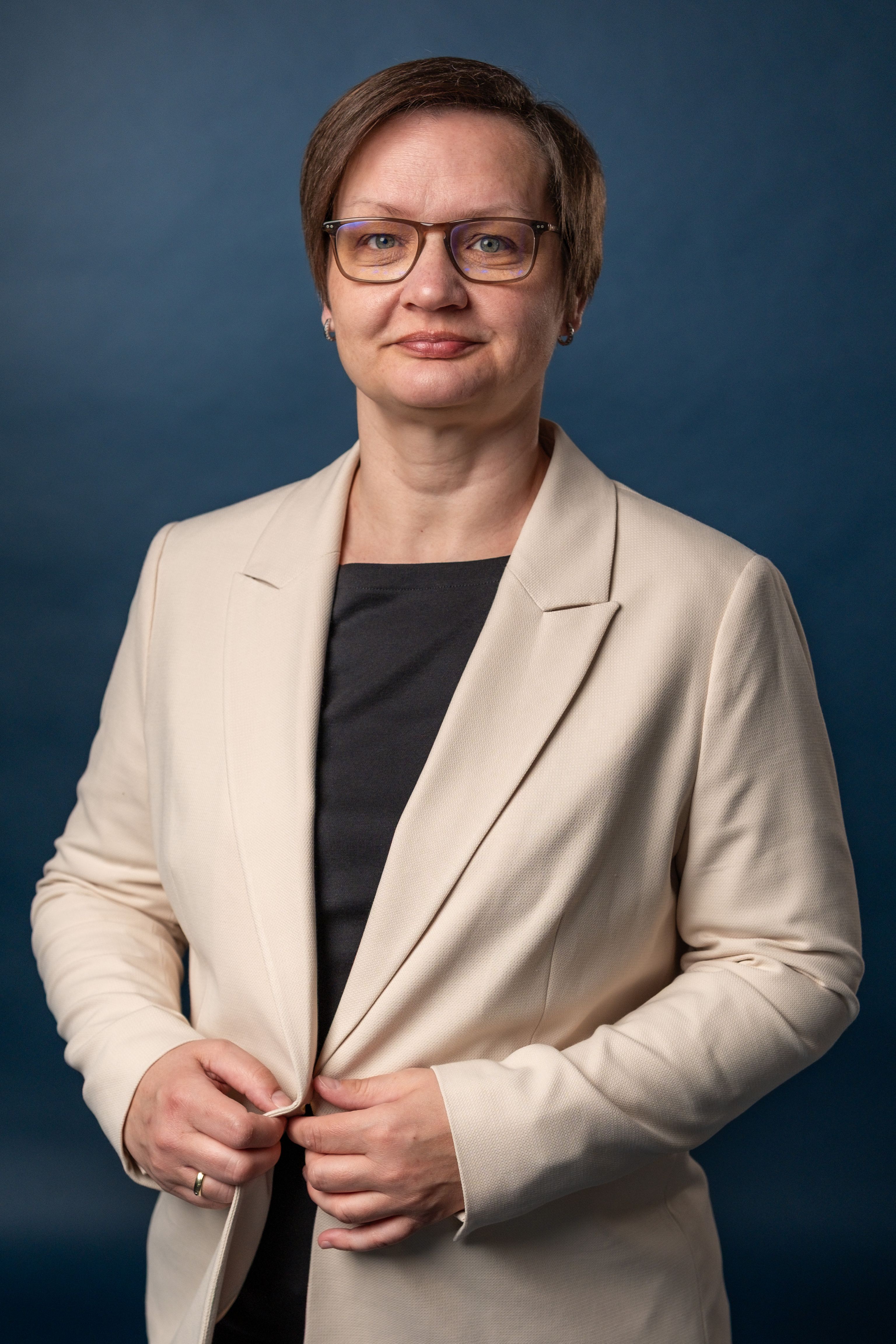 Frau mit Brille trägt beige Blazer über schwarzem Rollkragenpullover, lächelnd vor blauem Hintergrund.