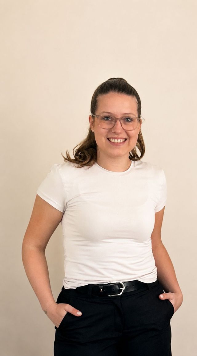 Lächelnde Person mit Brille in weißem T-Shirt und schwarzer Hose, Hände in den Hosentaschen.