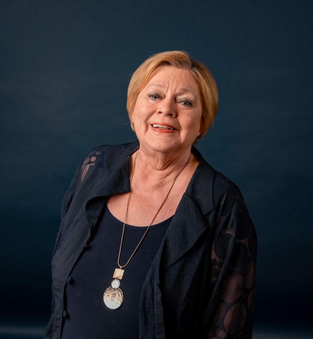 Lächelnde Frau mit schwarzem Blazer und goldener Medaille um den Hals vor dunklem Hintergrund.
