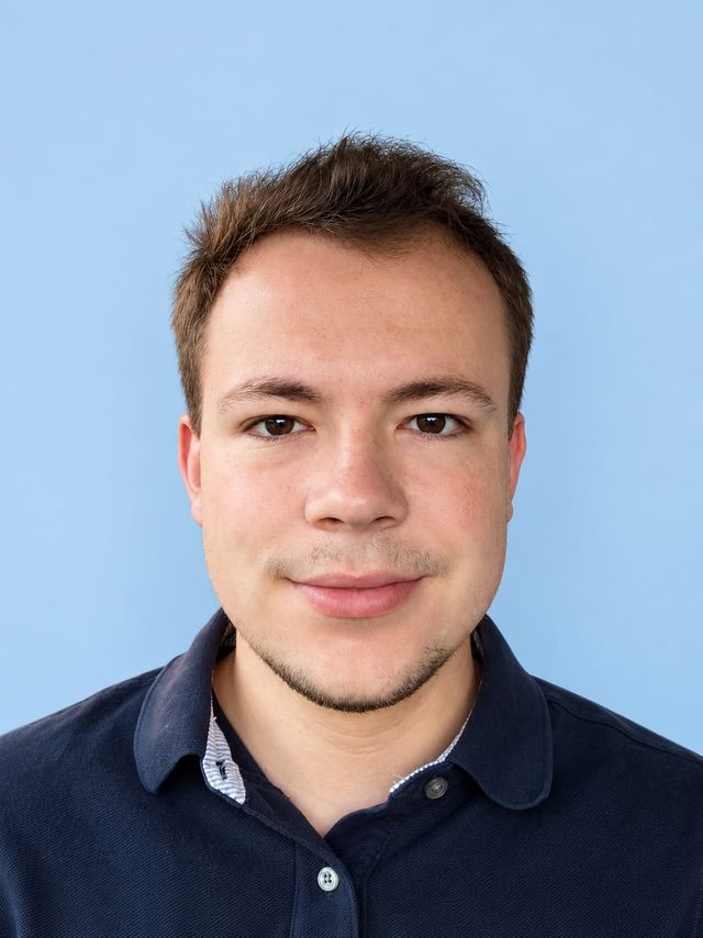Porträt eines lächelnden Mannes mit kurz geschnittenen Haaren und blauem Poloshirt vor hellem Hintergrund.