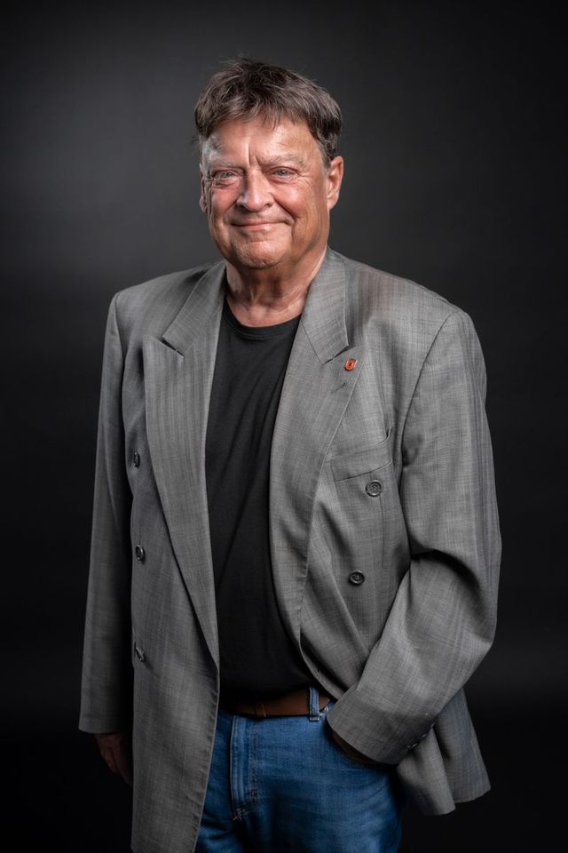 Lächelnder älterer Herr in grauer Jacke und schwarzem Shirt vor dunklem Hintergrund.